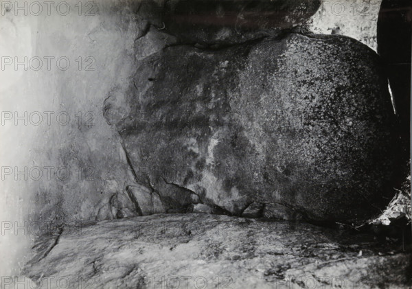 Keystone of the Kerguntuil dolmen, Trégastel, photograph, inscription: 'right - the keystone of the original, dolmen., Allée couverte de Kergantuil, Trégastel. C. du N.', photographer: attributed to Jan Lanting, supervision: Albert Egges van Giffen, 1939-05-27 - 1939-06-04, Attributed to, paper, baryta paper, gelatin silver print, developing gelatin silver print, height 123 mm x width 170 mm, megalithic chambered tomb, dolmen, A close-up view of a rough stone surface filling the frame, showing textured rock planes with cracks, crevices, and layered formations; areas of lighter and darker shading create mottled patterns, with one side appearing speckled and another side smoother; small fragments and chips of rock are visible along edges, and a narrow sliver of bright light or reflection appears near one edge; overall the image shows natural stone surfaces with varied textures and tones.