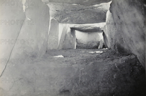 Kerguntuil Dolmen, Trégastel, photograph, inscription: 'afdr, Ban v He, Het interieur van de, allée couverte de Kergantuil, Trégastel C. du N., ZO richting', photographer: attributed to Jan Lanting, supervision: Albert Egges van Giffen, 1939-05-27 - 1939-06-04, Attributed to, paper, baryta paper, gelatin silver print, developing gelatin silver print, height 119 mm x width 170 mm, megalithic chambered tomb, dolmen, A low-angle, grayscale view of a narrow stone passage formed by large flat slabs serving as floor, side walls, and a horizontal capstone, with rough textured surfaces, scattered lighter patches on the ground, and bright light visible through the far end of the passage.