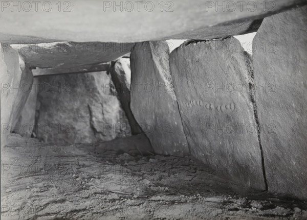 Kerguntuil Dolmen, Trégastel, photograph, inscription: 'Allée couverte de Kerganteul, Trégastel C. du N., carrying stone with the necklace (?), NW direction', photographer: attributed to Jan Lanting, supervision: Albert Egges van Giffen, 1939-05-27 - 1939-06-04, Attributed to, paper, baryta paper, gelatin silver print, developing gelatin silver print, height 123 mm x width 166 mm, megalithic chambered tomb, dolmen, A low, narrow space formed by large rough stone slabs with a textured dirt floor, the slabs showing visible striations and irregular surfaces, one vertical slab on the right featuring a row of shallow crescent indentations, light entering from a small gap near the top center and casting soft shadows along the interior, the ceiling slab appearing smooth compared to the sides, and scattered small debris and uneven patches across the ground.