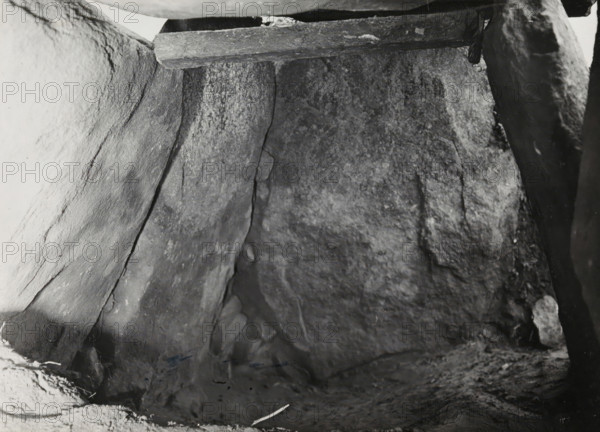 Kerguntuil Dolmen, Trégastel, photograph, inscription: 'the keystone seen from the inside, on the left the original corner filling, Allé couverte de Kerganteul, Tregastel. C. du Nord, afd, Ban dH...', photographer: attributed to Jan Lanting, supervision: Albert Egges van Giffen, 1939-05-27 - 1939-06-04, Attributed to, paper, baryta paper, gelatin silver print, developing gelatin silver print, height 122 mm x width 168 mm, megalithic chambered tomb, dolmen, A close view of rough stone surfaces forming an enclosed space, with large flat slabs leaning and meeting at angles, visible texture and cracks in the rock, a horizontal wooden beam resting near the top edge, small rounded stones tucked between slabs, and scattered dark and light patches across the uneven floor and walls.