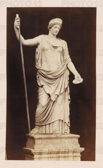 Sculpture of Juno, Museo Vaticano, photograph, photographer: Libreria Spithöver, c. 1850 - c. 1870, pasteboard, paper, albumen print, height 329 mm x width 216 mm, piece of sculpture, reproduction of a piece of sculpture, Vatican City, Vatican Museums, A full-length marble statue of a draped female figure standing on a stepped rectangular plinth is shown against a dark background, wearing a flowing, layered gown with detailed folds and a belted drapery at the waist, her left arm extended slightly forward holding a round, flat object, her right arm raised and grasping a long staff that reaches from the plinth to above her shoulder, her head turned slightly to one side and topped with a rounded headpiece, and her feet visible with sandals resting on the base.