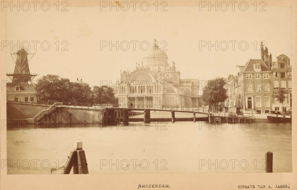 Amsterdam, View of the Palace of Popular Industry in Amsterdam, photograph, publisher: Andries Jager, c. 1865 - c. 1880, photographer, paper, pasteboard, albumen print, height 90 mm x width 145 mm, temporary exhibition, 'Salon', canal, façade (of house or building), A sepia-toned riverside scene showing a wide stretch of water in the foreground, a wooden post projecting from the water near the bottom edge, and a low arched bridge crossing the river toward the middle ground. Behind the bridge a large ornate building with a central dome and detailed arched windows dominates the center, flanked by trees on the left and a row of narrow multistory buildings with steep roofs on the right. A windmill structure is visible at the far left, and small boats and dock structures line the right bank near the row of buildings. The sky appears pale and empty above the scene.