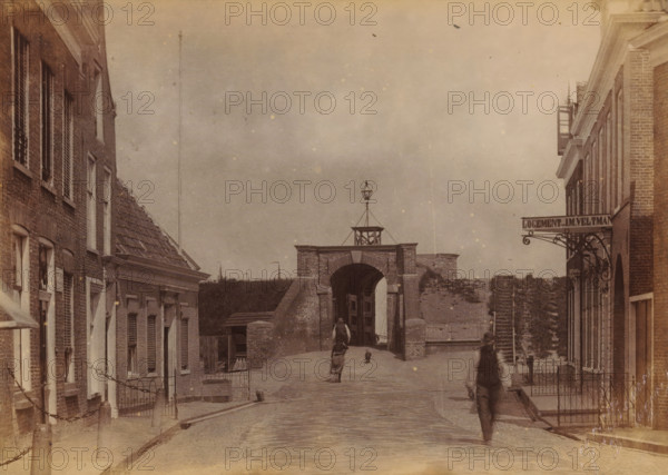 Street to the Great Water Gate in Delfzijl, photograph, name: 'JG Kramer', inscription: 'Poort van Delfzijl 20 Aug. 1895 prov Groningen', photographer: Johannes Gerardus Kramer, Delfzijl, 1895-08-20, paper, pasteboard, albumen print, albumendruk, height 98 mm x width 138 mm, street, city-gate, Grote Waterpoort, A sepia street scene framed by rows of brick buildings on either side with shuttered windows and decorative cornices, leading toward a brick archway topped by a small cupola and a lamp structure, a sloped cobblestone road rising to the arch, a few indistinct figures walking along the street with one figure closer to the foreground and another near the arch, low iron fences and stone posts along the sidewalks, and a plain sky above speckled with small blemishes.