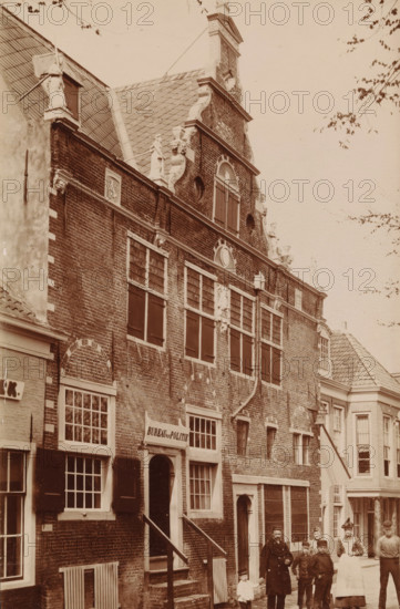 Facade of the Weigh House in Enkhuizen, photograph, inscription: 'Enkhuizen', Enkhuizen, c. 1885 - c. 1910, photographer, paper, pasteboard, daylight gelatin silver print, height 147 mm x width 97 mm, weigh-house, façade (of house or building), Waag (Enkhuizen), A brick building with stepped gables and decorative sculpted figures along the roofline fills most of the image, showing multiple shuttered windows and rectangular panes, arched brickwork above some windows, a tiled roof with a dormer, and ornamental stonework on the facade; a small flight of stairs leads to an arched doorway with a sign above it, and adjacent buildings with tiled roofs and sash windows are visible; a group of people stands on the street in front of the building, including an adult man holding a small child's hand, other children, and a woman wearing a long skirt and blouse, with branches and sparse leaves of a tree visible at the top and right edges of the scene.