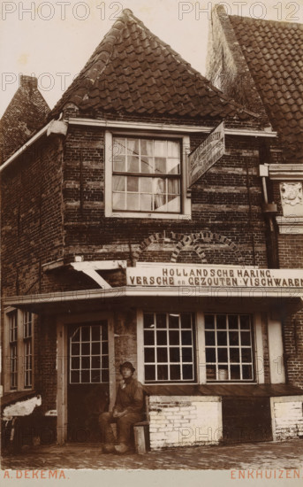 Exterior of a shop selling fish products and ship supplies in Enkhuizen, photograph, inscription: 'Enkhuizen', photographer: Albert Dekema, Enkhuizen, c. 1900, paper, pasteboard, daylight gelatin silver print, height 149 mm x width 97 mm, shop, store, façade (of house or building), A sepia photograph of a brick building with a steep tiled roof and a large upper window with panes and curtains, a small awning over a storefront below, and a man wearing a hat and work clothes seated on a small bench outside the entrance. The facade shows brickwork with decorative arches above the awning, a multi-paned display window next to a paneled door, a wooden support beam under the awning, and a carved decorative panel on the right side. The street in front is cobbled and there are worn bricks and a low raised ledge along the front of the building.