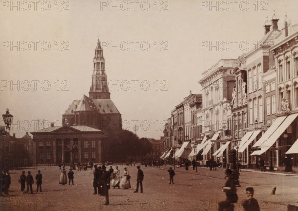 View of the Fish Market in Groningen, photograph, inscription: 'Groningen', photographer: Johannes Gerardus Kramer (possibly), Fish Market (Groningen), c. 1880 - c. 1900, Groningen (city), paper, pasteboard, albumen print, height 97 mm x width 137 mm, square, circus, church (exterior), A wide street scene with a large multi-tiered church tower rising behind a classical building with columns, a row of ornate buildings with awnings along the right side, and groups of people standing and walking across the open paved square; lamp posts appear near the foreground and along the street, and the sky above is clear and pale.