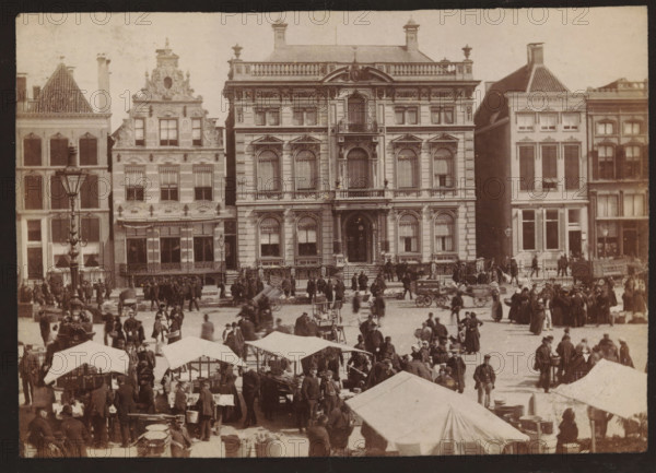 Buildings along the Grote Markt in Groningen, photograph, inscription: 'Groningen', name: 'JG Kramer Groningen', photographer: Johannes Gerardus Kramer, Grote Markt (Groningen), c. 1880 - c. 1900, Groningen (city), paper, pasteboard, albumen print, height 96 mm x width 139 mm, square, circus, façade (of house or building), A wide street scene shows a row of multi-story stone buildings with ornate facades and large windows, the central building featuring arched windows, decorative balconies, and a grand entrance with steps. In front of the buildings a busy open market fills the foreground with numerous canvas tents and stalls, carts, wooden crates, and barrels. Many people in period clothing stand, walk, or gather in small groups across the square, some near the stalls and others by horse-drawn carts. A tall, decorative lamppost stands near the left side of the image, and a few bicycles and handcarts are visible among the crowd. The overall tone of the photograph is sepia, with visible ...