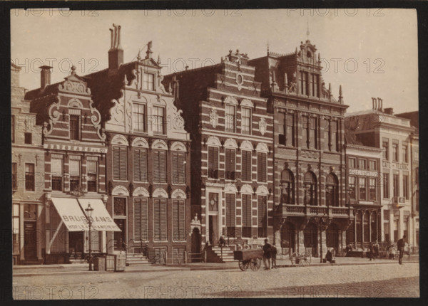 Buildings along the Grote Markt in Groningen, photograph, inscription: 'Groningen', photographer: Johannes Gerardus Kramer (possibly), Grote Markt (Groningen), c. 1880 - c. 1900, Groningen (city), paper, pasteboard, albumen print, height 96 mm x width 139 mm, square, circus, façade (of house or building), A row of ornate multi-story buildings with decorative facades and stepped gables lines a cobblestone street, each building featuring tall windows with shutters, arched doorways, and sculptural ornamentation on the upper levels; an awning extends from one storefront above the sidewalk, and wooden barrels and crates sit near the entrance; a few horse-drawn carts and a couple of horses stand on the street in front of the buildings, and small groups of people gather along the sidewalk and near doorways.