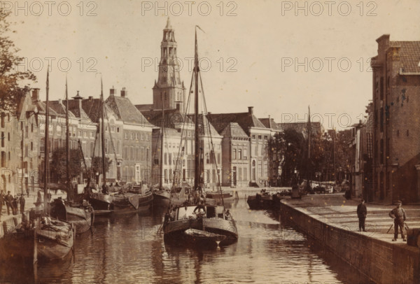 View of the Hoge der A in Groningen, with the tower of the Der Aa-kerk in the background, photograph, inscription: 'Groningen', photographer: Johannes Gerardus Kramer (possibly), Groningen (city), c. 1880 - c. 1900, paper, pasteboard, albumen print, albumendruk, height 96 mm x width 142 mm, canals, waters (in city), square, circus, ships (in general), A-kerk, A canal scene with several moored sailboats and small boats aligned along the water, tall masts rising above the vessels and reflections visible on the water surface, a row of multi-story brick buildings with pitched roofs and shuttered windows along the far side of the canal, a prominent tower rising above the buildings in the background, trees lining parts of the waterfront, a paved quay or walkway on the right side where a few people stand, additional boats and rigging along both sides of the canal, and stone walls edging the water.