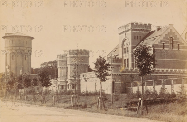 Exterior of the Cellular Penal Prison in Groningen, photograph, inscription: 'Groningen?', name: 'JG Kramer Groningen', photographer: Johannes Gerardus Kramer, Groningen (city), c. 1880 - c. 1900, paper, pasteboard, albumen print, height 96 mm x width 142 mm, façade (of house or building), prison, jail, Cellulaire Strafgevangenis, A sepia-toned street view shows a row of young trees with wooden supports along a grassy verge in front of a long brick wall topped with decorative stone battlements, behind which stand several brick buildings with castle-like architectural details including round towers with crenellated tops, a taller square tower with arched window openings, stepped gable ends, narrow vertical windows, and a large cylindrical water tower with a conical roof visible toward the left.