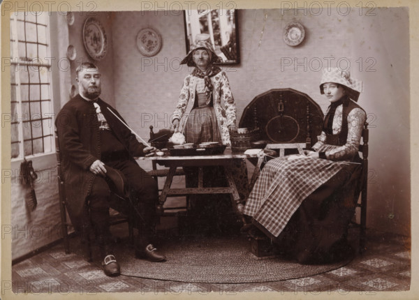 Portrait of an unknown man and two unknown women in traditional costumes from Hindeloopen, photograph, inscription: 'Th. M. Staas court photographer Leeuwarden Willemskade WL', inscription: 'Photograaf HJ de Koe Willemskade, W 1 Leeuwarden', photographer: Thomas Martin Staas, Hindeloopen, c. 1880 - c. 1900, paper, pasteboard, carbon print, height 99 mm x width 140 mm, family group, especially parents with their child(ren), folk costume, regional costume, interior, representation of a building, An indoor scene with three seated people around a low wooden table; a man in dark clothing with a beard sits on the left holding a hat and a long thin object, a woman in patterned traditional dress and a tall bonnet stands behind the table holding a teapot, and a young woman in patterned sleeves and a checked apron and bonnet sits on the right holding a book or paper on her lap; the table holds several bowls and a tray, and a basket and round wooden board lean against the wall behind them; the ro...