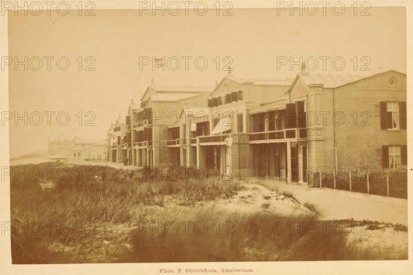 Facade of a building along the beach of Scheveningen, photograph, inscription: 'Scheveningen', photographer: Pieter Oosterhuis, Scheveningen, c. 1880 - c. 1900, paper, pasteboard, albumen print, height 94 mm x width 147 mm, beach, façade (of house or building), A sepia-toned photograph of a row of two-story buildings with balconies and shuttered windows facing a sandy, grassy foreground; some buildings have decorative pediments and finials on their roofs and one building displays a flag on a pole; a narrow path runs parallel to the buildings and a simple fence appears near one building; the foreground shows clumps of beach grass and patches of sand, and a distant shoreline is faintly visible.