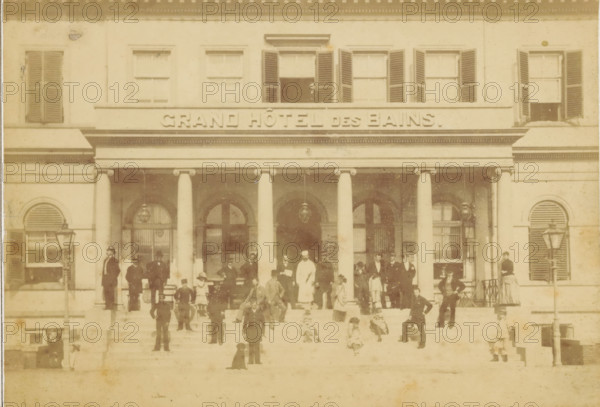 Group portrait of (presumably) employees on the steps of the Municipal Baths (Grand Hôtel des Bains) in Scheveningen, photograph, inscription: 'CP Wollrabe sen, The plate is kept until reordered.', photographer: Carl Philip Wollrabe, Scheveningen, c. 1870 - in or before 1884, paper, pasteboard, albumen print, height 101 mm x width 147 mm, working class, laborers, public baths, façade (of house or building), Grand Hôtel des Bains, A faded sepia photograph of a large building façade with a row of tall columns supporting a portico and a balcony above, multiple shuttered windows on the upper level, and arched doorways and windows behind the columns; a group of people in period clothing stand and sit on the wide front steps and under the portico, including men in suits and hats, women in long dresses, a person in a white uniform, children, and a seated dog; gas lamps flank the entrance, and decorative architectural details appear along the cornice and around the windows.