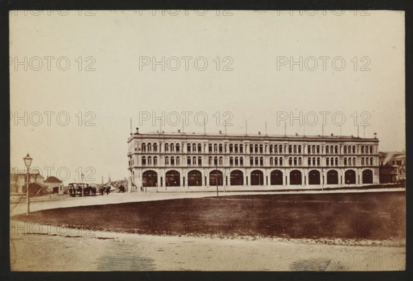 Side facade of a building in Scheveningen, photograph, inscription: 'CP Wollrabe sen, The plate will be kept until reordered.', photographer: Carl Philip Wollrabe, Scheveningen, c. 1870 - c. 1885, paper, pasteboard, albumen print, height 92 mm x width 142 mm, beach (as place of recreation), façade (of house or building), A wide, three-story stone building with a long row of arched openings on the ground level and evenly spaced arched windows on the upper levels stretches across the middle of the image, with thin vertical flagpoles projecting from its roof and decorative cornices along each floor; in front of the building there is an open paved area and a grassy expanse, and to the left of the building a cluster of smaller structures and a horse-drawn carriage with a few standing figures are visible near street lamps, while the sky above is pale and largely empty.
