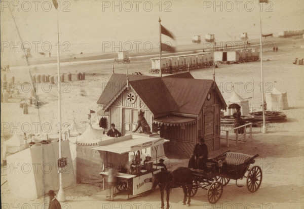 Wooden pavilion on the beach at Scheveningen, photograph, inscription: 'CP Wollrabe sen., The plate will be kept until reordered.', photographer: Carl Philip Wollrabe, Scheveningen, c. 1885 - c. 1900, paper, pasteboard, albumen print, height 100 mm x width 143 mm, beach (as place of recreation), four-wheeled, animal-drawn vehicle, eg: cab, carriage, coach, façade (of house or building), A sepia-toned beach scene shows a wooden pavilion with decorative trim and a peaked roof topped by a flag on a pole, a carved animal statue on a pedestal in front of the pavilion, and a man standing beside its entrance; in front of the pavilion is a cart with metal urns and a canopy, attached to a horse-drawn carriage with upholstered seating where a woman in dark dress and a hat sits, and another person stands nearby holding an umbrella; behind them are rows of small wheeled bathing huts on the sand and several tents with pointed tops, scattered people on the beach, piles of coiled material, and distan...