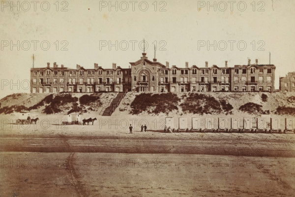 Exterior of the Oranje Hotel in Scheveningen, photograph, inscription: 'CP Wollrabe sen., The plate is kept until reordered.', photographer: Carl Philip Wollrabe, Scheveningen, c. 1885 - c. 1900, paper, pasteboard, albumen print, height 98 mm x width 146 mm, beach (as place of recreation), hotel, hostelry, inn, façade (of house or building), Oranjehotel, A wide, multi-story building with ornate architectural details stretches across a sandy embankment, centered by a taller central section with an arched feature and a narrow double staircase leading down the slope, flanked by lower wings with repeated windows and small balconies; grassy and shrub-covered dunes sit between the building and the beach, where a row of small rectangular beach carts on wheels lines the shore, several horses hitched to simple carts stand nearby, and a few people in period clothing are scattered along the sand, some standing and some near the carts.