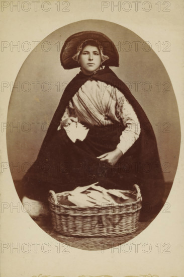 Portrait of an unknown woman in traditional costume, photograph, inscription: 'CP Wollrabe sen., The plate will be kept until reorder.', photographer: Carl Philip Wollrabe, South Holland, c. 1885 - c. 1900, paper, pasteboard, albumen print, height 149 mm x width 98 mm, adult woman, folk costume, regional costume, container made of plant material other than wood: basket, A sepia-toned portrait of a seated figure wearing a wide, rounded bonnet with ruffled edges and a dark cape draped over the shoulders, a patterned shawl visible across the chest and a long dark skirt covering the legs; one hand holds a folded piece of cloth while the other rests on the skirt, and a woven basket filled with folded linens sits on the floor in front of the figure against a plain oval background.