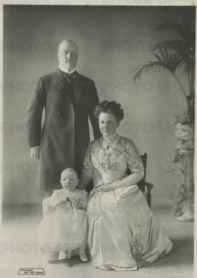 Portrait of Wilhelmina, Queen of the Netherlands, Hendrik of Mecklenburg-Schwerin and Juliana, photograph, Hendrik and Juliana. Bottom left 'ORIGINAL'., address: 'Guy de Coral & Co HOF-FOTOHANDEL AMSTERDAM-DEN HAAG.', photographer: Coral & Co., Guy de, Netherlands, 1909 - 1911, paper, pasteboard, photography, height 149 mm x width 106 mm, height 235 mm x width 146 mm, The title mentioned here refers to the highest title obtained by the person portrayed, not necessarily to the title the person portrayed bears in this image., Person, Hendrik (Prince of the Netherlands (1876-1934)), Juliana of the Netherlands, Wilhelmina of the Netherlands, A tall man in a dark formal coat stands behind a seated woman and a small child; the man has a moustache and wears a high-collared shirt with a tie. The woman sits on a wooden chair wearing a long, ornate dress with lace and embroidered details, a layered necklace, and her hair styled up; one hand rests on the child's shoulder and the other is in her l...