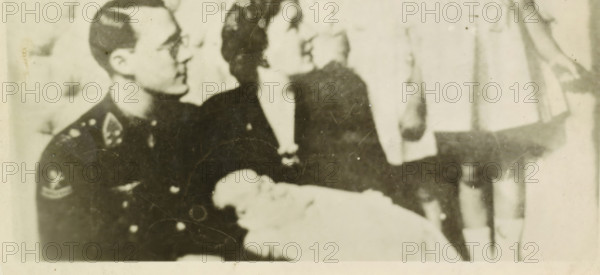 Portrait of Juliana, Queen of the Netherlands, Bernhard van Lippe-Biesterfeld, Beatrix, Irene, Princess of the Netherlands, and Margriet, photograph, Bernhard, Irene and Margriet in Ottawa., inscription: 'found in the field at Bocholtz L 1940-45 Signed H Dolmans 1980', Netherlands, 1943 - 1944, photographer, paper, photography, height 81 mm x width 113 mm, The title mentioned here refers to the highest title obtained by the person portrayed, not necessarily to the title the person portrayed bears in this representation., Person, Margriet (Princess of the Netherlands), Juliana of the Netherlands, North America, Ottawa, A grainy black-and-white photograph showing three seated figures and a bundled infant; on the left a person in a dark uniform with visible insignia on the shoulder and chest looks toward the center, wearing glasses and short hair; in the center a person with dark hair in a dark dress or coat faces slightly to the right, their face turned upward; on the right a lighter-ton...