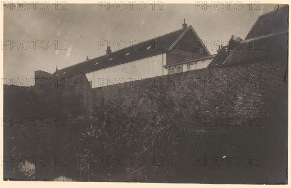 Part of the city wall of Harderwijk, photograph, annotation: 'g, Harderwijk', annotation: 'City wall, Harderwijk, 1900 - 1920, photographer, Attributed to, paper, pasteboard, gelatin silver print, height 136 mm x width 217 mm, city-walls, A low viewpoint shows a tall brick wall running across the image with foliage visible in the foreground; behind the wall a long building with a steep tiled roof and several chimneys rises, one gable end with a triangular peak and a window opening is visible, and a portion of a sloped tiled roof on the right has a seated figure near the ridge; the sky above is plain and slightly mottled.