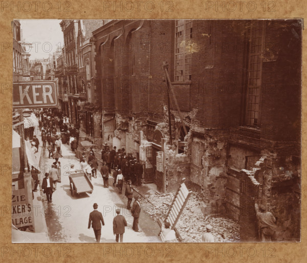 Street view with the Nieuwezijds Chapel in Amsterdam during the demolition in 1908 (former title), View of the Kalverstraat with the Nieuwezijds Chapel in Amsterdam during the demolition in 1908, photograph, annotation: ''08.150, 446', annotation: 'NZ Chapel', Amsterdam, 1908, photographer, paper, pasteboard, daylight collodion silver print, height 118 mm x width 154 mm, height 369 mm x width 295 mm, street (city(-scape) with figures, staffage), church (exterior) - QQ - small church, chapel, Kalverstraat, Nieuwezijds Chapel, A sepia street scene framed by tall brick buildings shows a busy thoroughfare with many pedestrians wearing period clothing and hats, some carrying umbrellas and others pushing a handcart. The left side features shopfronts and awnings lining the narrow street while the right side displays a large, weathered brick wall with tall windows and exposed, crumbled lower masonry revealing rubble and construction debris. A group of people congregates near a doorway set into...