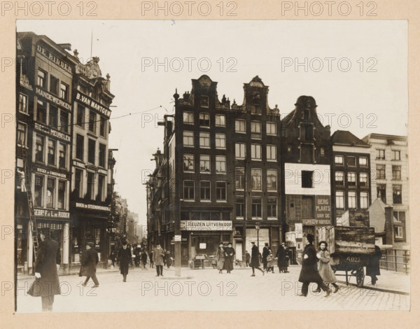View of Sophiaplein or Muntplein in Amsterdam, photograph, annotation: ''17.2, Gift from photographer C. Steenbergh, Sophiaplein, On the right 5 houses being demolished', annotation: 'Sophiaplein (= Muntplein)', photographer: Cornelis Johannes Steenbergh (possibly), Amsterdam, 1900 - 1917, Cornelis Johannes Steenbergh, photographer, paper, pasteboard, gelatin silver print, developed gelatin silver print, height 126 mm x width 172 mm, height 183 mm x width 237 mm, square, circus, etc. (city(-scape) with figures, staffage), Muntplein (Amsterdam), A street scene with rows of tall narrow buildings featuring ornate gables and many windows, a mix of dark and light façades, and shopfronts at ground level; people in period clothing walk and stand on a cobbled square, some wearing coats and hats, one person pulling a small cart, a horse-drawn wagon with a visible number parked near stacked wooden crates, and a few lampposts and a central pole with overhead wires stretching between buildings.