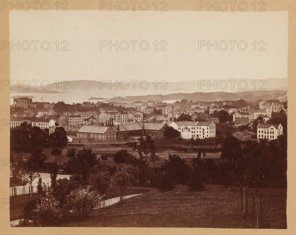 View of Christiania, Christiania, photograph, photographer: Olaf Martin Peder Væring, 1881, pasteboard, paper, albumen print, albumendruk, height 145 mm x width 183 mm, height 245 mm x width 330 mm, city-view in general, 'veduta', A sepia-toned panorama showing a townscape with rows of multi-story buildings and houses set among trees and open grassy areas, a large rectangular industrial-looking building with a sloped roof and chimneys near the center, clusters of residential structures with varied rooflines and windows spread across a gently rolling landscape, a foreground of grassy fields and scattered young trees with protective stakes and a winding dirt path, a body of water and low-lying islands visible beyond the town, and distant hazy hills forming the horizon under a pale sky.