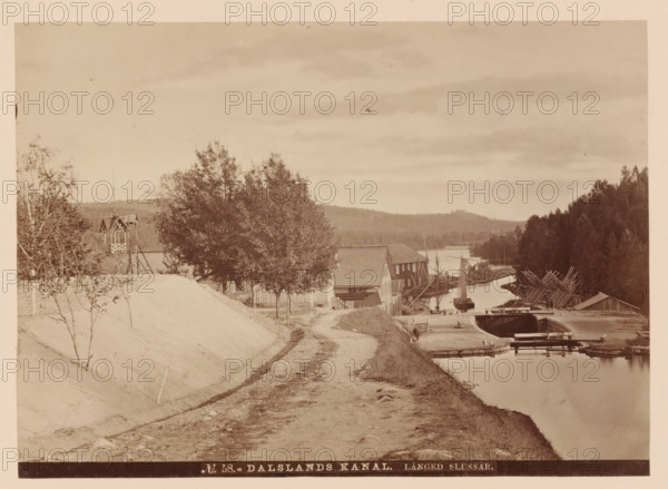 Lock in the Dalsland Canal in Sweden, Dalsland kanal, långed slussar, photograph, number: 'No 58.a', retailer's mark: 'FR. HULDBERG, Bazarin å Norrbro', photographer: Robert Dahllöf, Göteborg, 1872 - 1874, Robert Dahllöf, photographer, pasteboard, paper, albumen print, albumendruk, height 147 mm x width 205 mm, height 233 mm x width 310 mm, canal, sluice, weir, A sepia-toned riverside scene shows a dirt path running alongside a narrow canal with calm reflective water, wooden buildings and boathouses lining the far bank, a small sailboat moored near a dock, piles of long logs stacked along the shoreline, and several trees clustered near the buildings and on the embankment, with a forested hill visible in the distance under a cloudy sky.