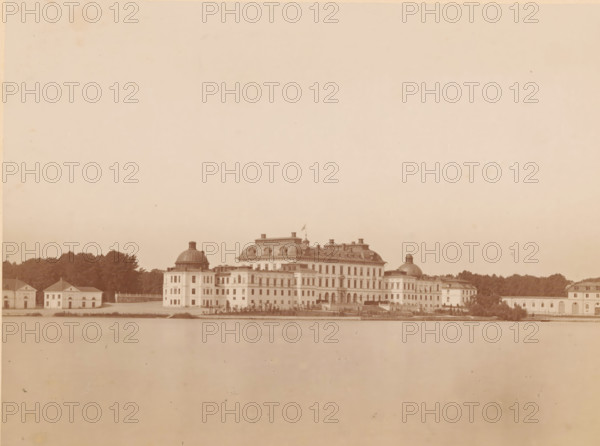 View of Drottningholm Castle near Stockholm, Drottningholms Slott, photograph, photographer: August Roesler, Stockholm, 1862 - 1893, pasteboard, paper, albumen print, albumendruk, height 161 mm x width 220 mm, height 268 mm x width 320 mm, palace, Drottningholm Castle (Ekerö), Ekerö, A wide sepia-toned photograph shows a large classical building complex along a waterfront, composed of a central grand structure with a mansard roof and ornamental rooftop elements flanked by domed pavilions and lower rectangular wings, with multiple rows of windows and arched openings on the ground level; a flag flies atop a small tower on the central roof. The complex sits behind a low waterfront embankment and a narrow strip of water in the foreground, and is set against a backdrop of dense trees and smaller outbuildings at each end. The sky above is expansive and pale, occupying most of the image, and the overall scene has an aged, uniform brownish tone with slight spots and blemishes on the surface.