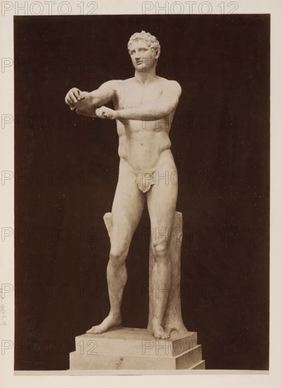 Apoxyomenos, Sculpture of Apoxyomenos, Vatican, album page, photograph, This photo is part of an album., inscription: '1315 M. Vat. APOXYOMENOS', photographer: attributed to James Anderson, Vatican Museums, after sculpture by anonymous, c. 1857 - c. 1875, after sculpture by, Vatican City, Attributed to, paper, albumen print, height 382 mm x width 273 mm, piece of sculpture, reproduction of a piece of sculpture, athletics, gymnastics, A full-length photograph of a marble male statue standing on a stepped rectangular base against a dark background, showing a nude figure with short curly hair and a calm facial expression, the torso turned slightly with both arms extended forward and hands positioned as if holding something, one forearm crossed over the other, a cloth draped over the groin, muscular legs with one foot placed forward and the other slightly back, a tree-trunk-like support behind one leg, and a smooth stone plinth beneath the feet.