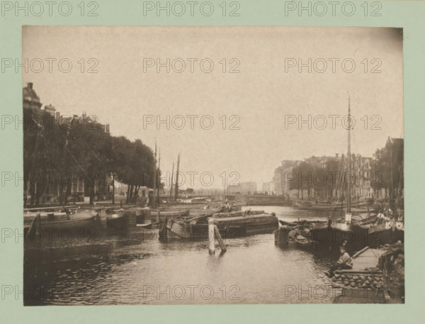 View of Amsterdam, album page, photomechanical print, This print is part of an album., inscription: 'Mij. voor Photolithographr & Zincographie, procédé Mr. EJ Asser.', Amsterdam, printer (person): Maatschappij voor Photolithographie & Zincographie, 1892 - c. 1900, printer (person), paper, collotype, photogravure, height 170 mm x width 234 mm, city-view in general, 'veduta', A sepia-toned waterfront scene showing a canal lined with moored boats and barges, masts rising along both sides, trees and multi-story buildings along the water's edge, a low bridge in the distance spanning the canal, wooden posts and docks extending into the water, rippled reflections on the canal surface, and a person seated on a dock near stacked coiled ropes at the lower right.