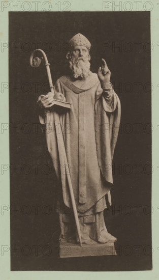 Image of a bishop, album page, photomechanical print, This print is part of an album., inscription: 'Mij. voor Photolithographr & Zincographie, procédé Mr. EJ Asser.', printer (person): Maatschappij voor Photolithographie & Zincographie, Amsterdam, 1892 - 1900, printer (person), paper, collotype, photogravure, height 281 mm x width 153 mm, archbishop, bishop, etc. (Roman Catholic), A monochrome photograph of a full-length statue of a robed figure wearing a mitre and a long flowing robe, with a long beard and mustache; the figure holds a curved staff in one hand and a book with a round object on it in the other, while the opposite hand is raised with two fingers extended; the robe features layered drapery and decorative tassels at the hem, and the statue stands on a rectangular plinth against a dark background with a pale border surrounding the image.
