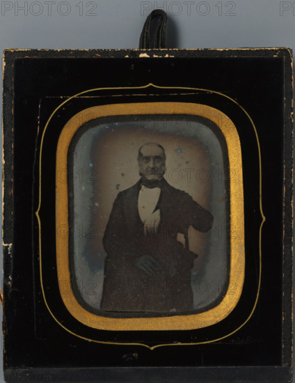 Portrait of Wouter Holleman, manufacturer, photograph, inscription: 'Wouter, 1831.', Netherlands, c. 1855 - c. 1870, photographer, pasteboard, inorganic material, glass, paper, ambrotype, height 74 mm x width 58 mm, height 129 mm x width 111 mm, Person, Wouter Holleman, historical persons, A framed photographic image shows an older man seated and facing forward, wearing a dark suit jacket with a white shirt and bow tie, one arm resting on the arm of a chair and the other hand visible near his lap; the photograph is surrounded by a decorative mat with an inner gold oval border and an outer black frame with gold trim, and a looped black cord is visible at the top of the frame.
