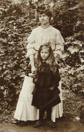 grün No 25 m. Fou ? auflage., gray at 6 fach Block, Portrait of a woman and a girl in front of a bush, photograph, annotation: 'grün No 25 m. Fou ? auflage., ... gray im 6 fach Block', photographer: attributed to Richard Tepe, Netherlands, c. 1900 - c. 1930, Attributed to, pasteboard, paper, daylight gelatin silver print, height 153 mm x width 98 mm, A sepia-toned photograph showing a woman standing behind a young girl in an outdoor leafy setting, the woman wearing a light-colored blouse with decorative smocking and voluminous sleeves and a long light skirt while the girl wears a dark dress with long sleeves and a layered skirt; the woman's hands rest gently on the girl's shoulders and the girl has long loose hair with a bow at the top, both facing forward with neutral expressions, surrounded by dense foliage and undergrowth, and both visible from head to toe with shoes on a dirt path.