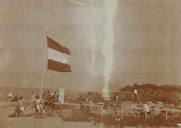 Beach at Valkeveen with the Dutch flag, photograph, inscription: 'o', Valkeveen, c. 1925 - c. 1950, photographer, paper, baryta paper, daylight collodion silver print, height 91 mm x width 119 mm, beach (as place of recreation), flag, colors (as symbol of the state, etc.), A tall flag on a pole with horizontal dark light and dark bands flutters above a sandy beach crowded with people and beach chairs, a bicycle leaning near the pole, groups of seated and standing figures wearing hats and summer clothing, rows of folding chairs and small tables in the foreground, a rocky tree-covered headland at the shoreline in the distance, and a pale vertical streak running down the center of the image.