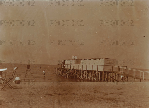 Beach at Valkeveen with changing rooms on the pier, photograph, A camera on a tripod can be seen on the beach on the left., inscription: 'Q', Valkeveen, c. 1925 - c. 1940, photographer, paper, baryta paper, daylight collodion silver print, height 91 mm x width 119 mm, beach (as place of recreation), pier, quay, wharf, A sepia-toned beach scene with a wooden pier structure extending over the water, built on a lattice of stilts and supporting a row of small changing cabins; a few people are on the pier and on the beach, some standing in shallow water and one person crouched on the sand; folding chairs and camera stands are positioned on the shore; the horizon shows calm water under a wide, empty sky.