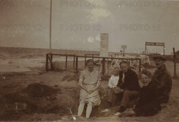 Portrait of five unknown people on the beach of Valkeveen, photograph, inscription: 'V', Valkeveen, c. 1925 - c. 1932, photographer, paper, baryta paper, daylight collodion silver print, height 59 mm x width 90 mm, beach (as place of recreation), A sepia-toned beach scene with a wooden pier extending into the sea and waves along the shoreline, worn sand and scattered seaweed in the foreground, a tall vertical post near the pier, a freestanding signboard on the beach, stacked deck chairs and a simple fence behind the group, and five people seated on the sand near the water: a woman in a patterned dress with folded hands, a small child leaning toward an older man in a dark jacket, a man wearing a cap and a coat, and another child with an open-mouthed expression.