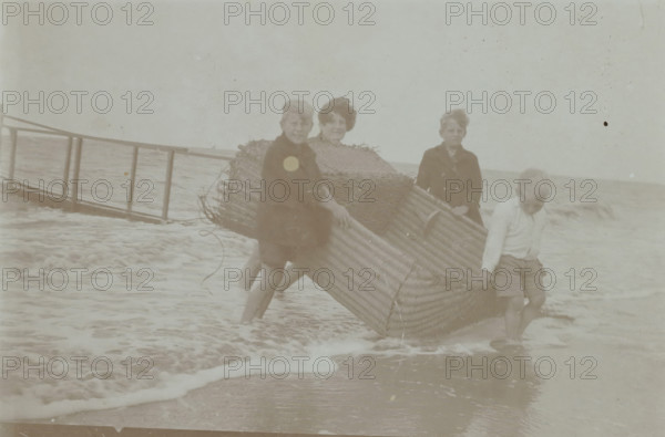 Koosje, Daan and Ben Ochse and another boy carrying a wicker beach chair out of the sea, photograph, inscription: 'AA', Valkeveen, c. 1925 - c. 1935, photographer, paper, baryta paper, daylight collodion silver print, height 59 mm x width 90 mm, Person, Koosje Ochse, Daniël Bernardus 'Ben' Ochse, Heinrich Daniël 'Daan' Ochse, beach (as place of recreation), child (four persons), A faded black and white scene at a shoreline shows four children standing in shallow water while holding a large woven wicker boat or basket. Three children face the camera, two standing close together with one partially behind the wicker, and a boy further right holding the edge and looking slightly down. Another child at the front right leans forward gripping the wicker and standing with wet legs. In the background a simple wooden railing or pier extends into the water, and small waves wash around the children and the base of the wicker, creating foamy water and wet sand at the foreground. The sky appears pal...