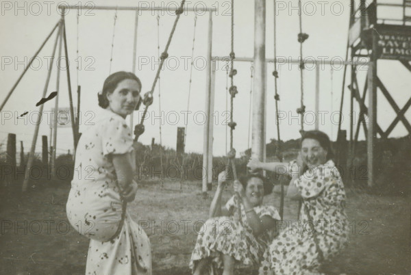 Three women on swings on the beach at Valkeveen, photograph, inscription: 'AM', inscription: '1936', Valkeveen, c. 1936, photographer, paper, baryta paper, gelatin silver print, height 65 mm x width 87 mm, beach (as place of recreation), playground, A faded black-and-white scene of three women on a playground swing structure, two seated on swings and one sitting sideways on a swing facing the camera, all wearing patterned dresses; the woman on the left looks toward the camera, the two on the right are smiling, and one of them holds a vertical pole; behind them are metal swing frames, vertical ropes with knots, grassy ground, wooden posts, and a tall metal lattice structure on the right.