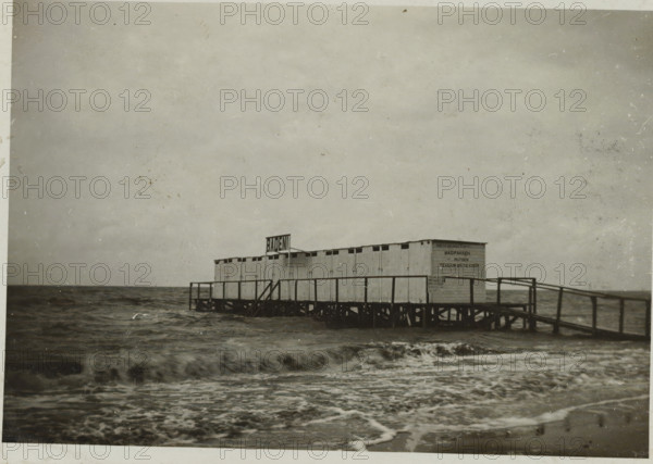 Pier with changing rooms in the Zuiderzee at the beach of Valkeveen, photograph, inscription: 'AI', Valkeveen, c. 1925 - c. 1950, photographer, paper, baryta paper, gelatin silver print, developed gelatin silver print, height 58 mm x width 82 mm, pier, quay, wharf, Zuiderzee, A long wooden pier structure with small connected cabin-like units stands on stilts over choppy dark water, a simple railing running along the pier and a set of stairs leading down toward the sea, waves breaking in the foreground and a wide expanse of cloudy sky above.
