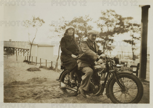 Wilhelm Ochse and Catharina Maria Groennou on a motorcycle on the beach of Valkeveen, photograph, inscription: '1934', inscription: 'AG', inscription: '4', Valkeveen, c. 1934, photographer, paper, baryta paper, gelatin silver print, developed gelatin silver print, height 60 mm x width 86 mm, Person, Catharina Maria Groennou, Wilhelm Ochse, beach (as place of recreation), motorcycles and other two-wheeled motor vehicles, A sepia-toned photo shows two people seated on an old motorcycle on a sandy surface near a wooden building and a raised pier, with trees and sparse vegetation in the background; the person in front wears a flat cap and a coat and has one hand on the handlebar while the person behind wears a cloche-style hat and a long coat and holds onto the seat, the motorcycle's visible parts include a front wheel with spokes, a headlamp, an engine, and a license plate mounted at the front, and a vertical wooden post appears at the right edge of the image.