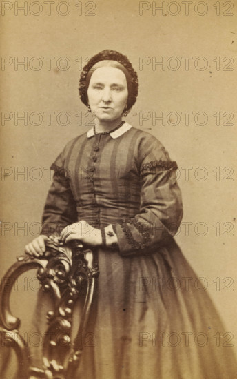 Portrait of a woman from the Marmelstein family, presumably the mother of AF Marmelstein, photograph, carte-de-visite, address: 'A. Greiner, Photograf, Nieuwendijk. L. 87, Amsterdam.', annotation: 'Grandmother Marmelstein.', photographer: Albert Greiner, Amsterdam, 1861 - 1874, paper, pasteboard, albumen print, albumendruk, height 105 mm x width 61 mm, historical persons not known by name - BB - woman, A sepia-toned portrait of a woman standing and resting her hands on the carved back of a wooden chair, wearing a dark dress with vertical stripes and decorative trim on the sleeves, a light collar, and a fitted bodice with buttons down the front; her hair is covered by a dark bonnet or cap, she wears earrings and a necklace, and the background is plain.