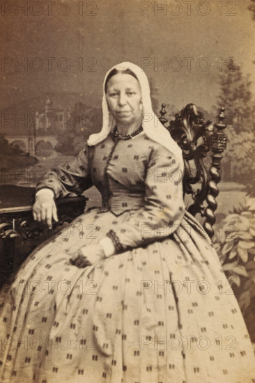 Portrait of an unknown woman from the Marmelstein family, photograph, carte-de-visite, photographer: Jacques Pilartz, Amsterdam, 1850 - 1900, paper, pasteboard, albumen print, albumendruk, height 101 mm x width 62 mm, historical persons, adult woman, A seated woman wearing a patterned dress with a fitted bodice and full skirt and a plain headscarf that frames her face sits in an ornate carved wooden chair; one hand rests on a table with carved details while the other rests in her lap, and a painted backdrop showing architectural and landscape elements is visible behind her.