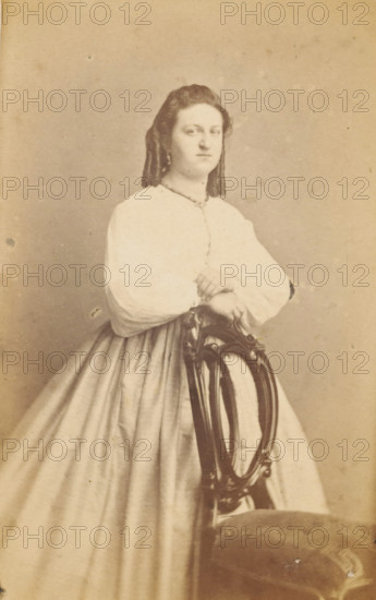 Portrait of an unknown woman from the Marmelstein family, photograph, carte-de-visite, inscription: 'J. KORSTEN, Painter, and PHOTOGRAPH, MIDDELBURG.', photographer: J. Korsten, Middelburg, 1864 - 1878, paper, pasteboard, albumen print, albumendruk, height 103 mm x width 61 mm, historical persons - BB - woman, A monochrome portrait of a woman standing and leaning with her folded hands on the back of an ornate wooden chair, wearing a light-colored dress with a full skirt and long sleeves, her hair styled away from her face and falling in waves, a neutral facial expression, and a plain backdrop behind her.