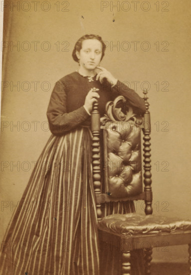 Portrait of an unknown woman from the Marmelstein family, photograph, carte-de-visite, photographer: Caspel & Stapert, S. van, Amsterdam, 1850 - 1900, paper, pasteboard, albumen print, albumendruk, height 110 mm x width 60 mm, historical persons, adult woman, A sepia-toned portrait of a woman standing beside an ornate wooden chair, wearing a dark bodice and a long skirt with vertical stripes; she rests one hand on a turned chair post and the other near her face, her hair pulled back, and the chair features a padded, tufted back and seat with carved decorative elements.