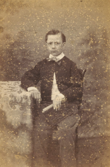 Portrait of an unknown boy from the Marmelstein family, photograph, carte-de-visite, address: 'PHOTOGRAPHISCH ATELIER., J. WIJNMAN, Halsteeg No. 248., Amsterdam.', photographer: J. Wijnman, Amsterdam, 1866 - 1870, paper, pasteboard, albumen print, albumendruk, height 107 mm x width 64 mm, historical persons not known by name, child, A sepia-toned portrait of a young person standing and facing the camera, wearing a dark jacket over a light shirt with a bow at the collar, one hand resting on a patterned cloth-covered surface and the other hand holding a rectangular object, with a plain studio backdrop and speckled marks across the image.