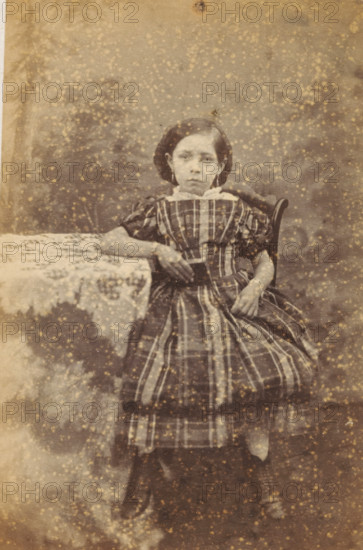 Portrait of an unknown girl from the Marmelstein family, photograph, carte-de-visite, address: 'PHOTOGRAPHISCH ATELIER., J. WIJNMAN, Halsteeg No. 248., Amsterdam.', photographer: J. Wijnman, Amsterdam, 1866 - 1870, paper, pasteboard, albumen print, albumendruk, height 108 mm x width 64 mm, historical persons not known by name - BB - woman, child, A seated young person with a solemn facial expression wears a striped dress with puffed sleeves and a white collar, one arm resting on a lace-covered table and the other hand holding the skirt; dark hair is styled close to the head and the figure sits on a wooden chair with a curved back, against a mottled backdrop with numerous light speckles and age spots visible across the image.