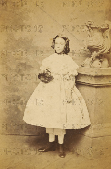 Portrait of an unknown girl from the Marmelstein family, photograph, carte-de-visite, address: 'ENGELSCH, Photographisch Etablissement, Kalverstraat bij de Munt, D. 39.', photographer: Engelsch Photographisch Etablissement, Amsterdam, c. 1865, paper, pasteboard, albumen print, albumendruk, height 98 mm x width 62 mm, historical persons not known by name - BB - woman, child, A young girl stands facing forward wearing a light-colored dress with puffed sleeves and a wide skirt, her dark hair styled in ringlets at the sides and parted in the middle, a dark choker at her neck, and dark shoes visible beneath a layer of light-colored underskirt; she holds a dark object near her waist with one hand while the other rests against a stone pedestal topped by an ornate sculpted urn with curved decorative elements, all set against a mottled studio backdrop.