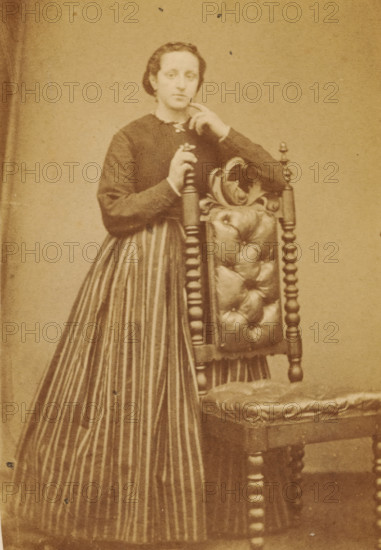 Portrait of an unknown woman from the Marmelstein family, photograph, carte-de-visite, photographer: Caspel & Stapert, S. van, Amsterdam, 1850 - 1900, paper, pasteboard, albumen print, albumendruk, height 110 mm x width 65 mm, historical persons, adult woman, A sepia-toned photograph shows a person standing beside an ornate wooden chair with a tufted upholstered back and seat; the person wears a dark long-sleeved bodice with a light collar and a long skirt with vertical stripes, has hair pulled back, and rests one hand on the top of the chair while the other hand is raised near the face.
