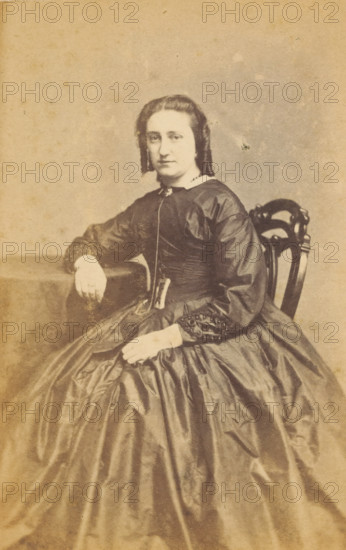 Portrait of an unknown woman from the Marmelstein family, photograph, carte-de-visite, photographer: J. Korsten, Amsterdam, 1850 - 1900, paper, pasteboard, albumen print, albumendruk, height 102 mm x width 60 mm, historical persons, adult woman, A sepia-toned portrait of a seated woman wearing a dark, full-skirted dress with a fitted bodice and long sleeves, her hair styled in center-parted waves that fall to about chin length; she sits on an ornately carved wooden chair, one arm resting on a covered table and the other hand placed on her skirt, and she wears gloves and a small decorative brooch at her collar.